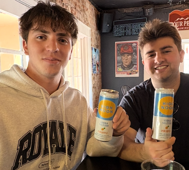 Two friends holding High Noon drinks inside a venue